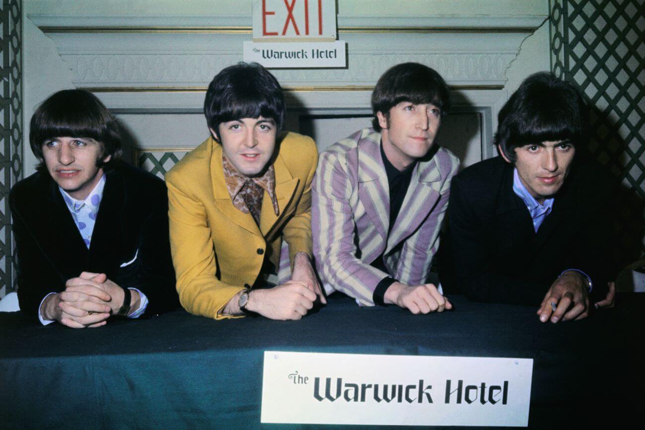 Ringo Starr, Paul McCartney, John Lennon, and George Harrison lean over a table in front of a door.