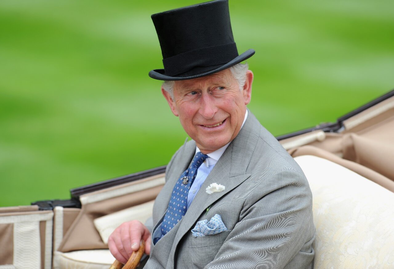 King Charles wears a top hat and smiles while riding in a horse and carriage.