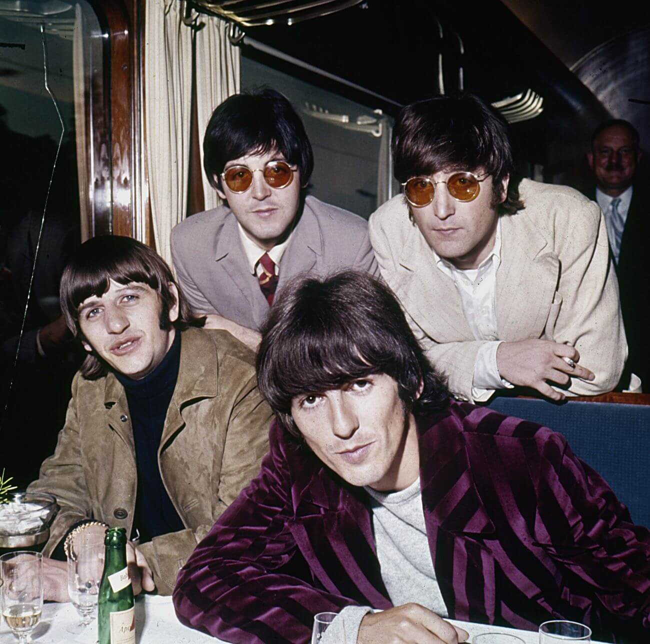 Ringo Starr, Paul McCartney, George Harrison, and John Lennon of The Beatles gather on one side of a table. McCartney and Lennon wear sunglasses.