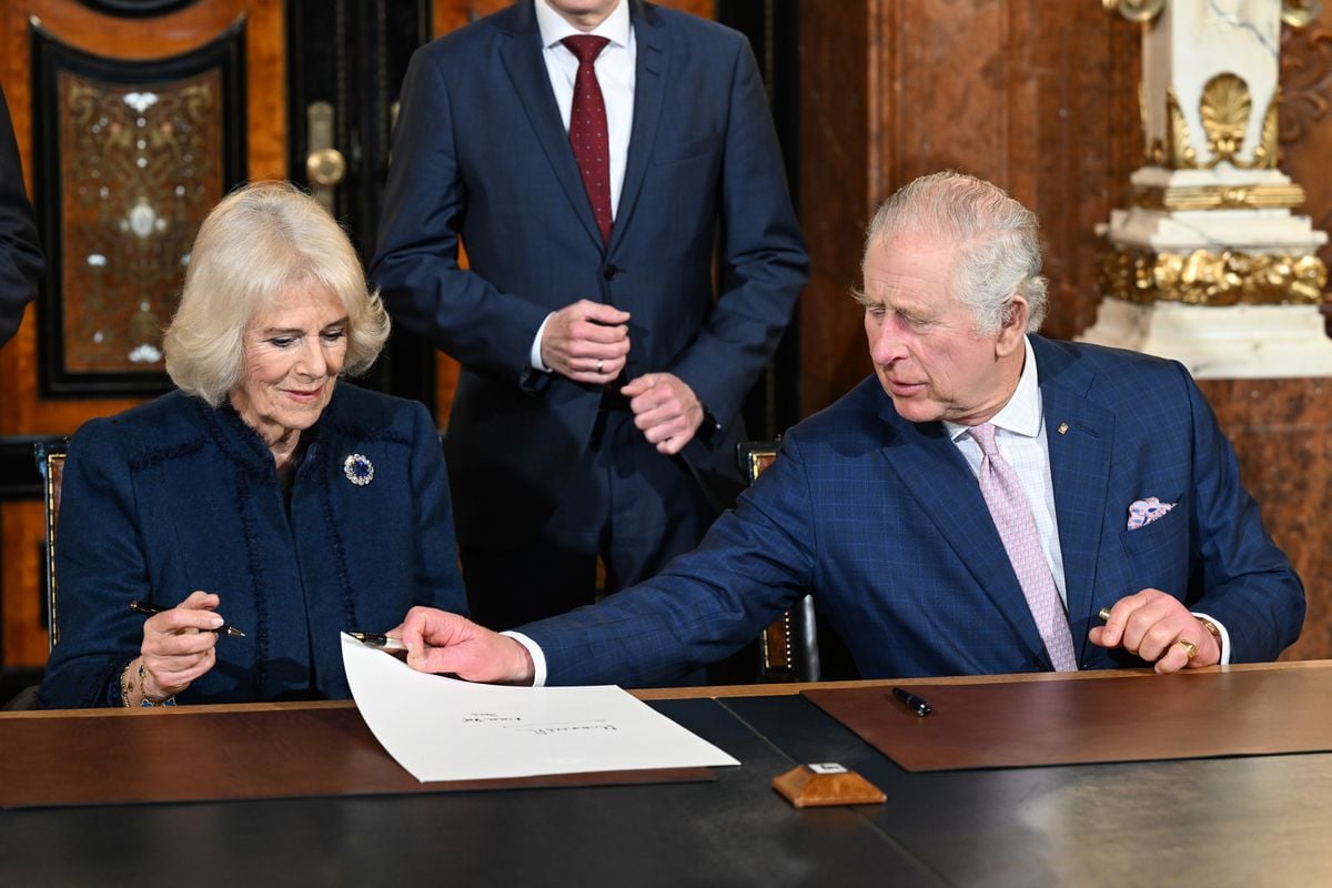 King Charles III and Camilla, Queen Consort sit together at a table signing a piece of paper together.