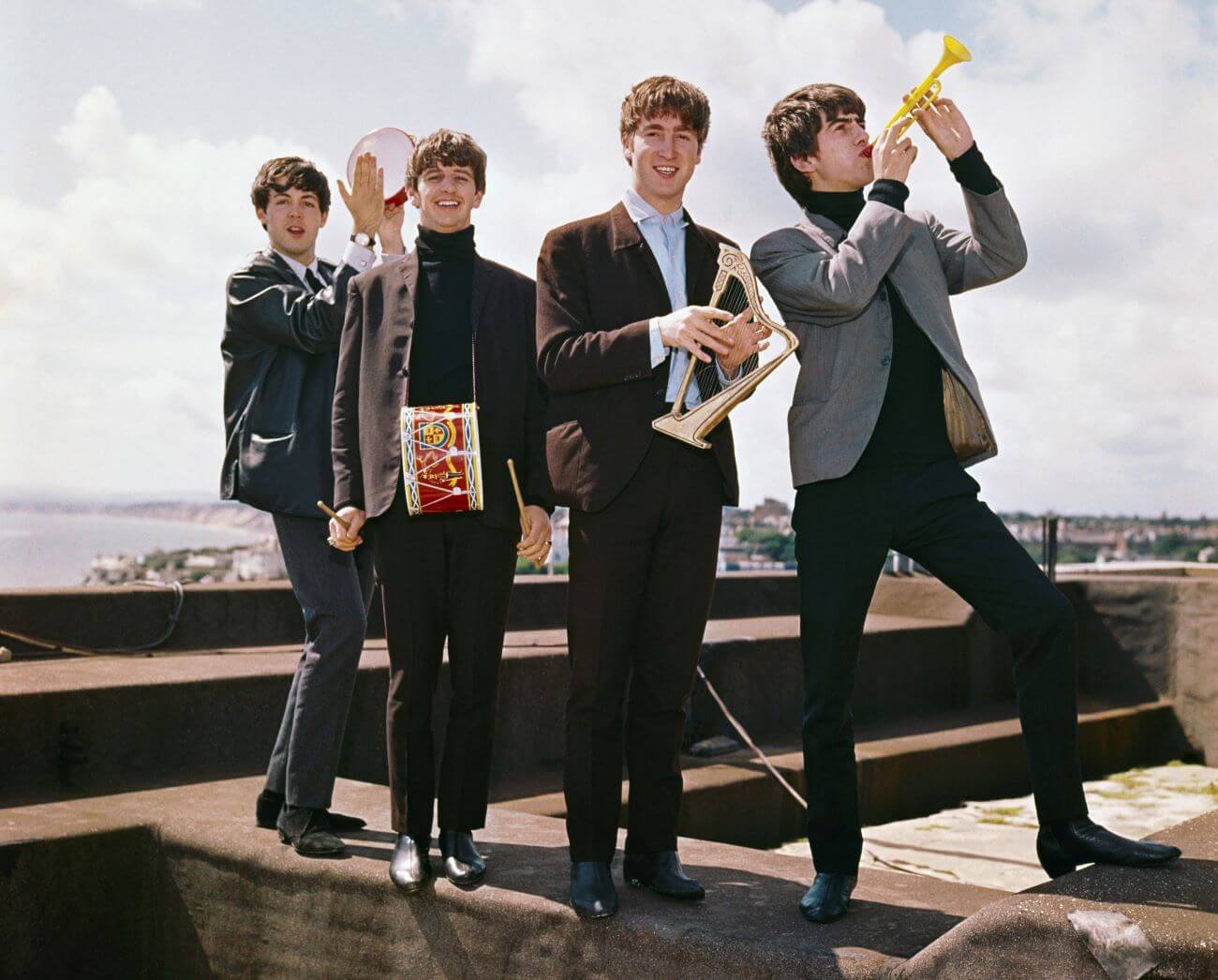 Paul McCartney, Ringo Starr, John Lennon, and George Harrison of The Beatles stand on a rooftop and play small instruments.