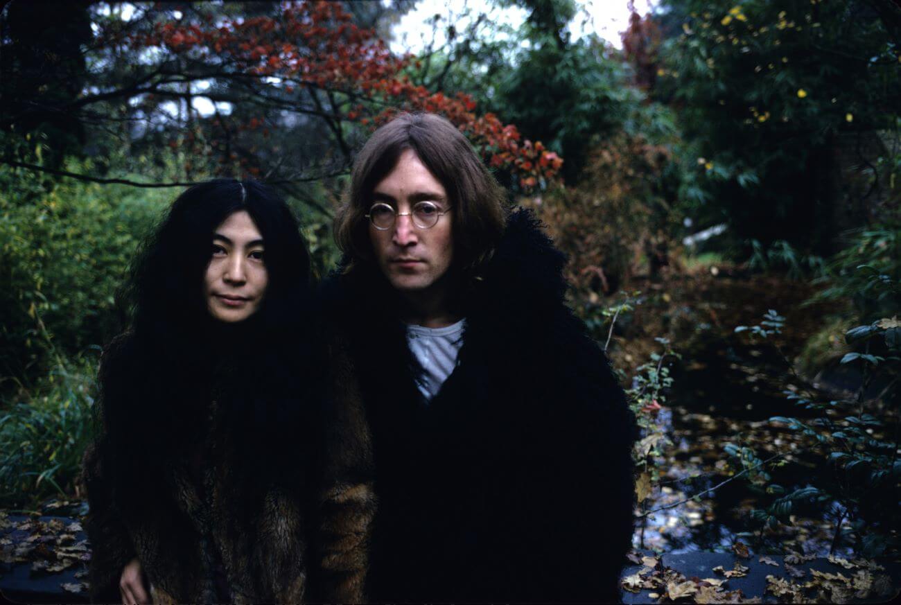 Yoko Ono and John Lennon wear black and stand in front of a forest backdrop.