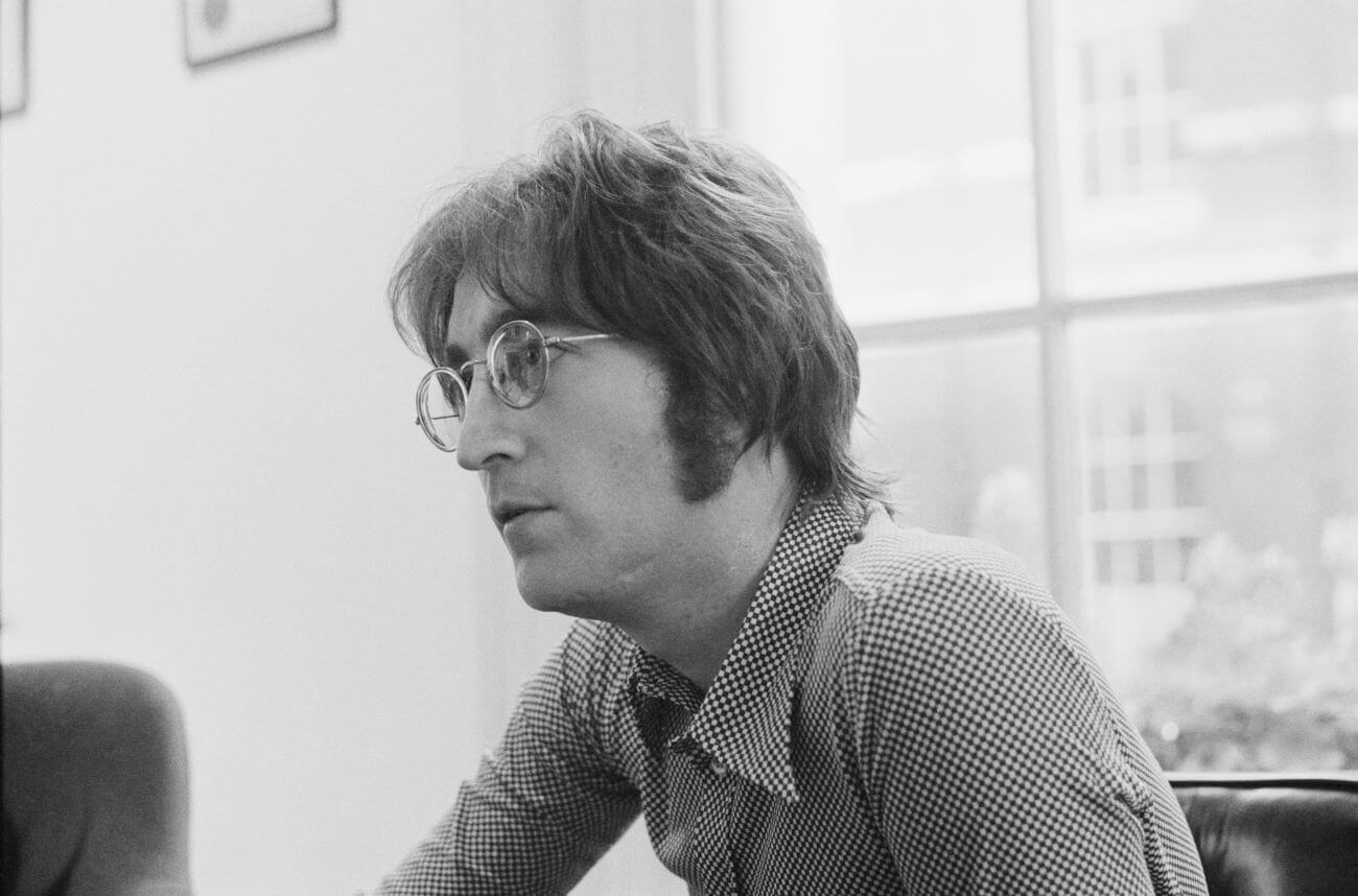 A black and white picture of John Lennon wearing a checked shirt and sitting by a window.