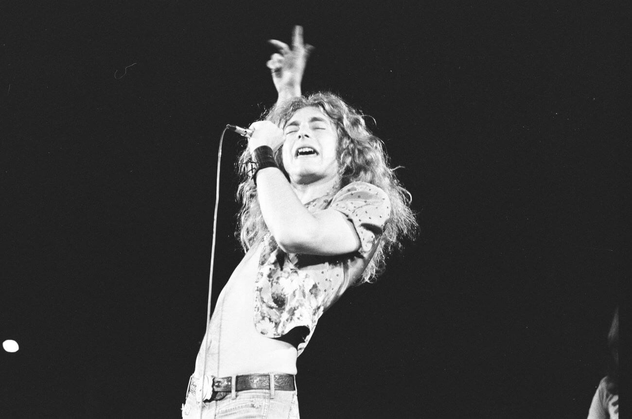 A black and white picture of Robert Plant singing into a microphone and holding one arm above his head.