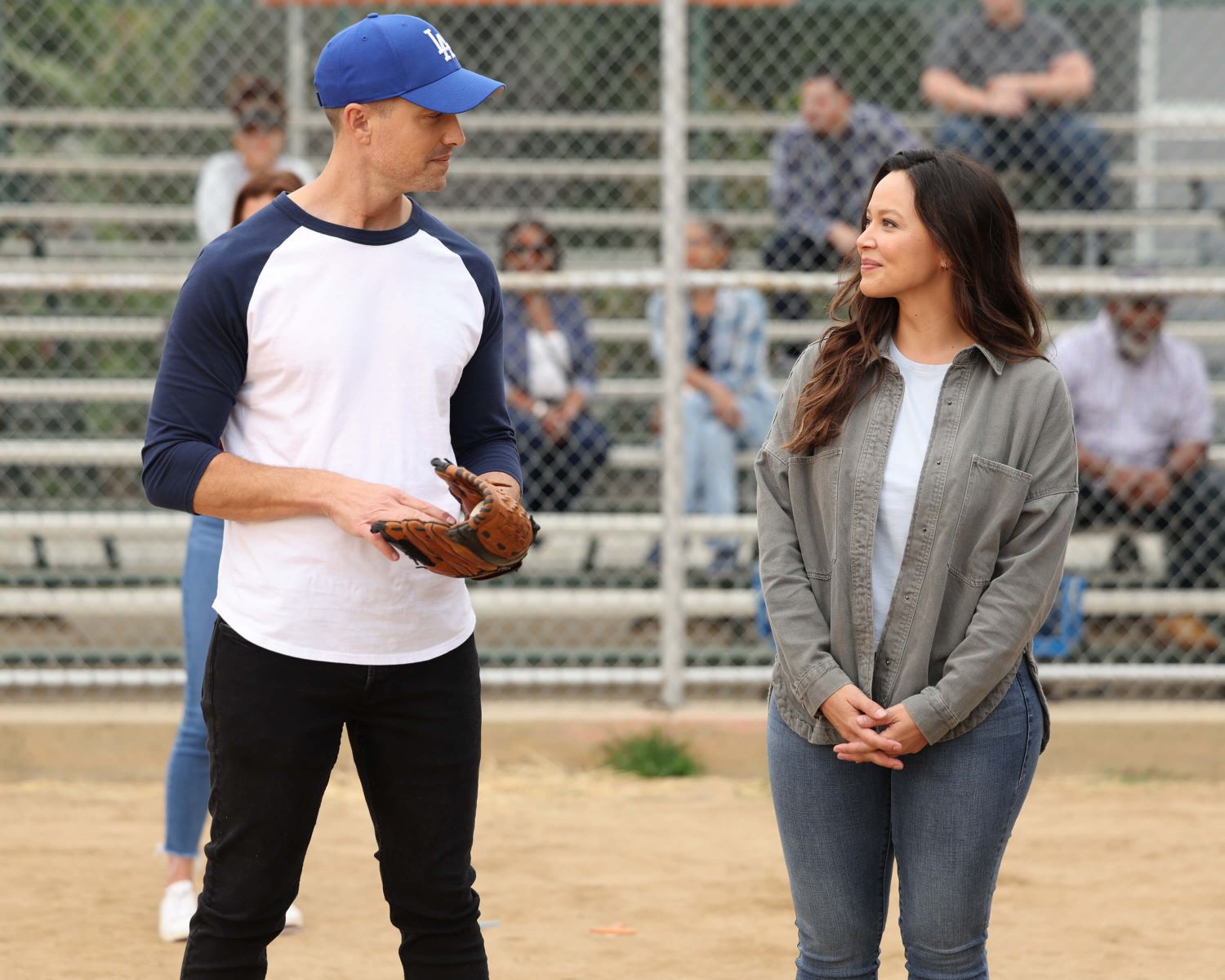 Eric Winter as Tim Bradford and Melissa O'Neil as Lucy Chen share a scene in 'The Rookie' Season 5 Episode 11. Tim wears a blue and white baseball shirt, black pants, and a blue Dodgers baseball hat. Lucy wears a gray button-up shirt over a white shirt and blue jeans.