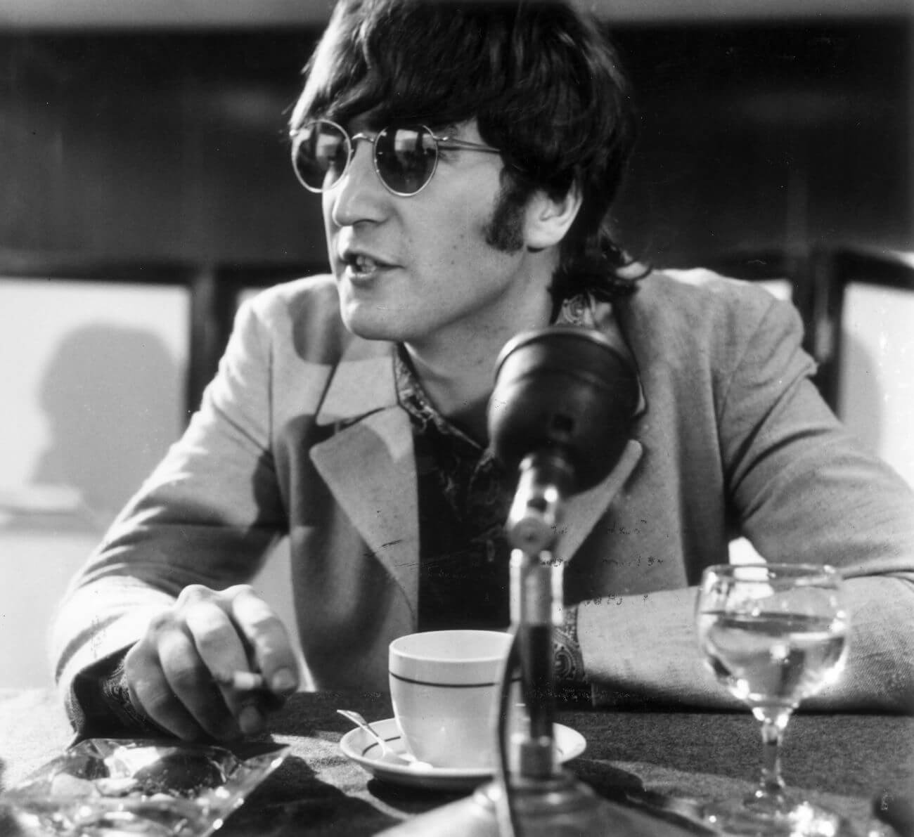 John Lennon of The Beatles wears sunglasses and sits in front of a microphone.