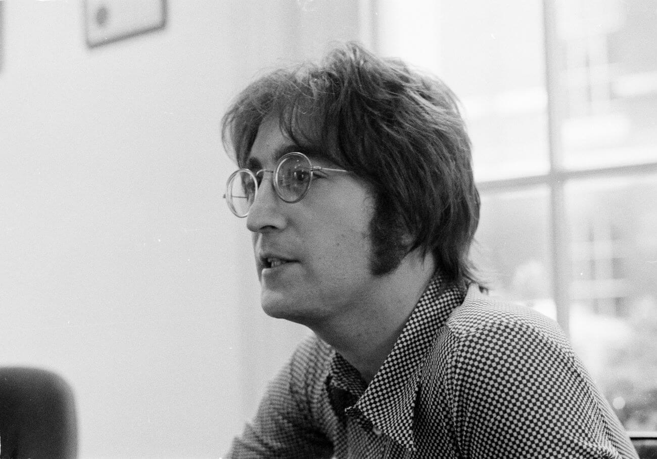 A black and white picture of John Lennon wearing a checked shirt and sitting in front of a window. 