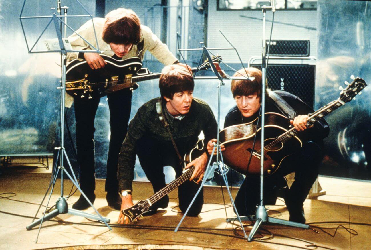 George Harrison, Paul McCartney, and John Lennon of The Beatles hold guitars and crouch on a stage.