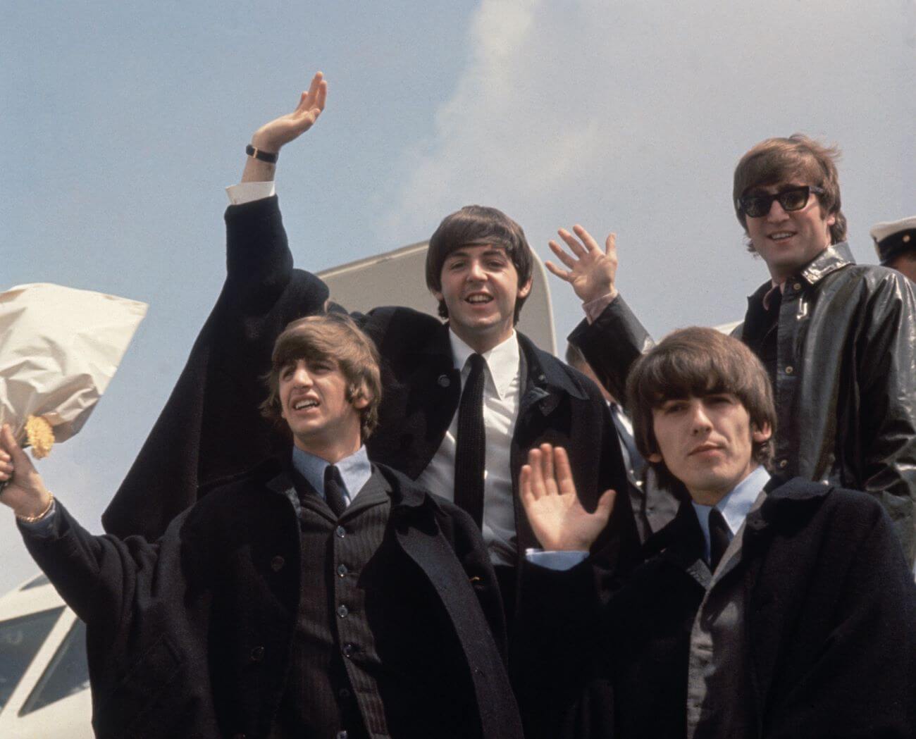 Ringo Starr, Paul McCartney, George Harrison, and John Lennon wave from the open door of an airplane.