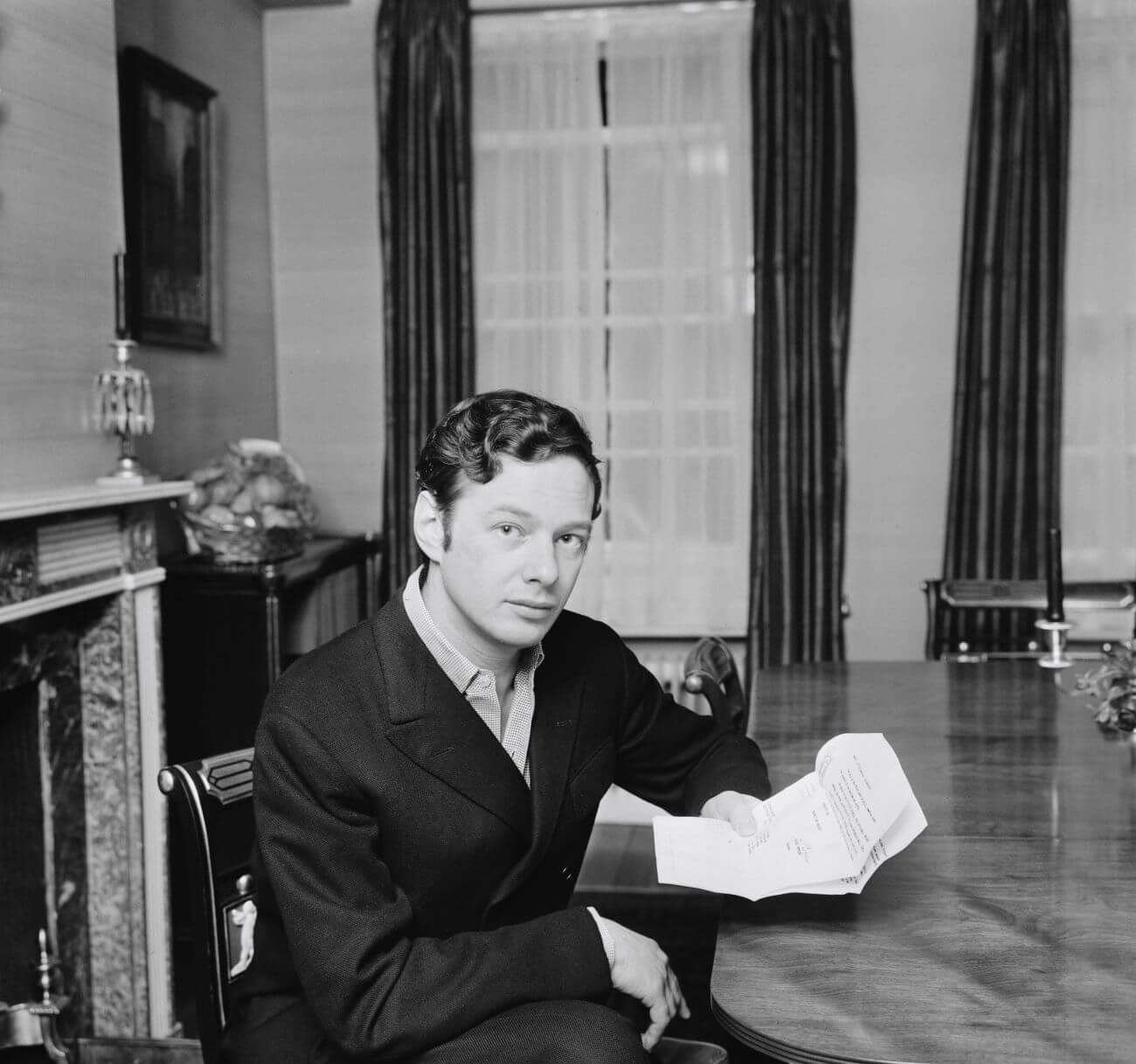 A black and white picture of Beatles manager Brian Epstein sitting a table holding papers.