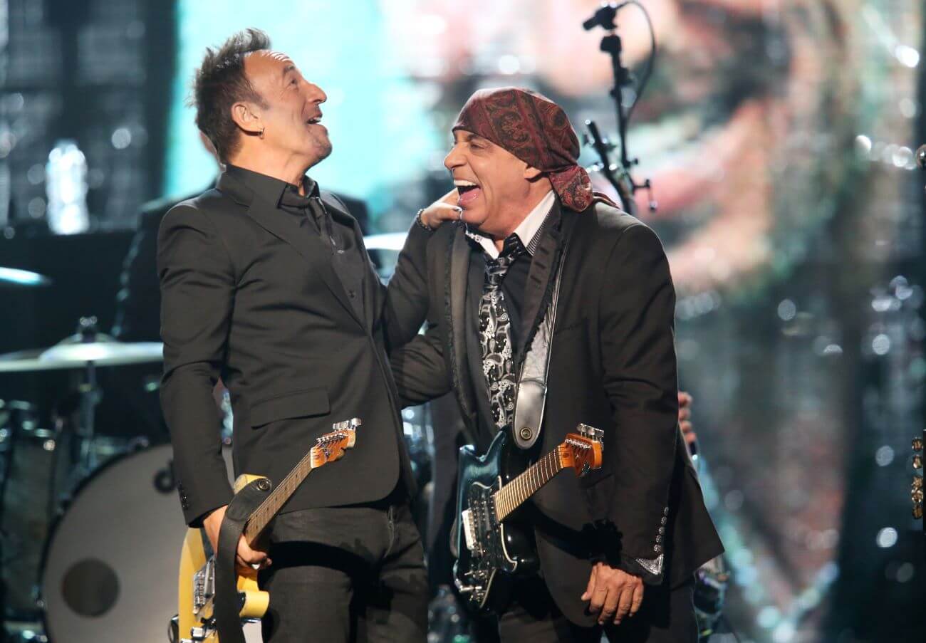 Bruce Springsteen grasps Steven Van Zandt's shoulder while they laugh onstage together.