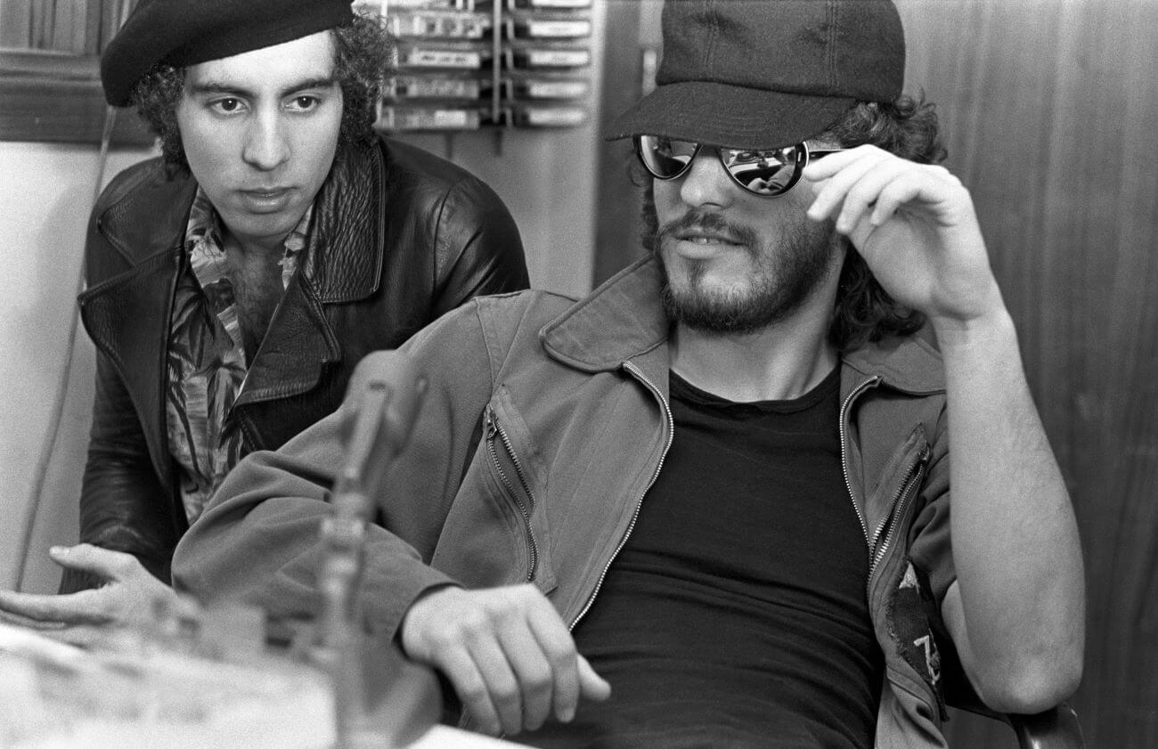 A black and white picture of Steven Van Zandt wearing a leather jacket and Bruce Springsteen wearing a baseball hat and sunglasses. They sit at a desk behind a microphone.