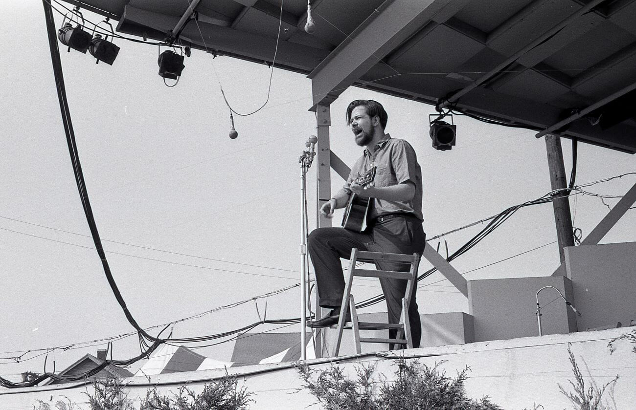 A black and white picture of Dave Van Ronk playing guitar and singing into a microphone. He rests one foot on a chair.