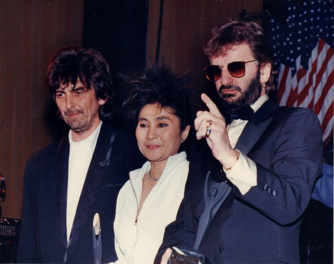 George Harrison, Yoko Ono, and Ringo Starr stand in front of an American flag. Starr wears sunglasses 