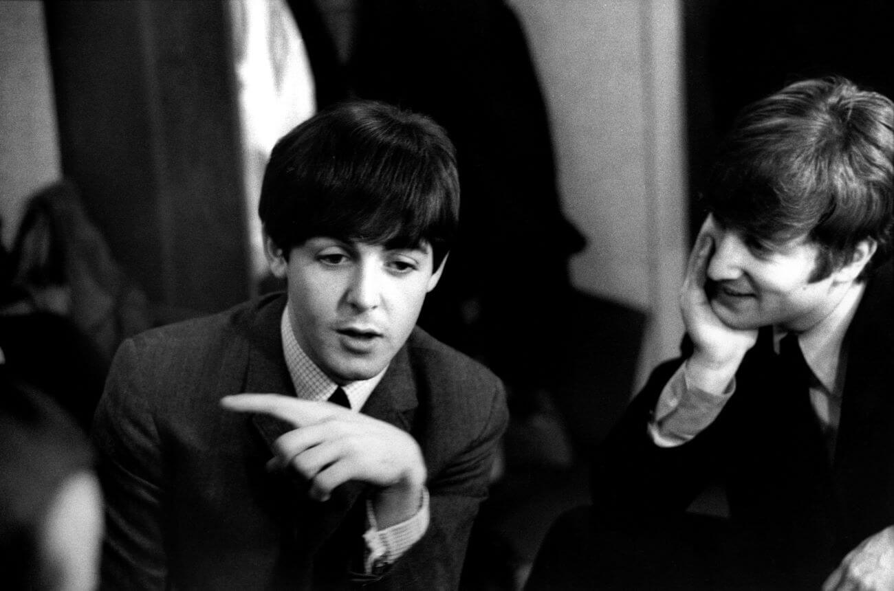 A black and white picture of John Lennon resting his chin in his hand and smiling while watching Paul McCartney talk.