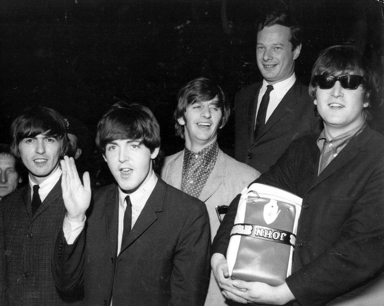 A black and white picture of George Harrison, Paul McCartney, Ringo Starr, Brian Epstein, and John Lennon. Lennon wears sunglasses and holds a bag.