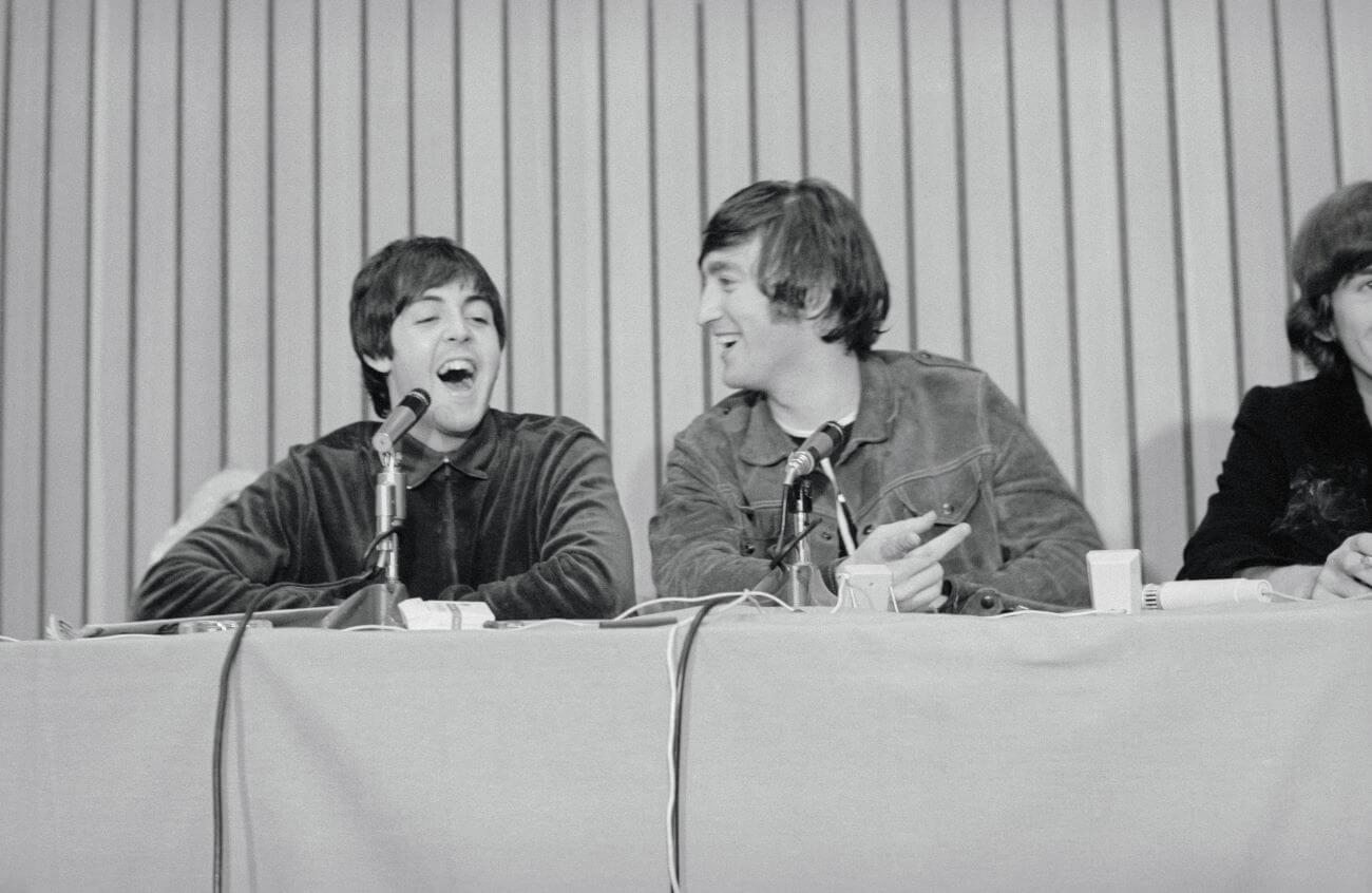 A black and white picture of Paul McCartney and John Lennon sitting at a table together laughing. A microphone is in front of both of them.