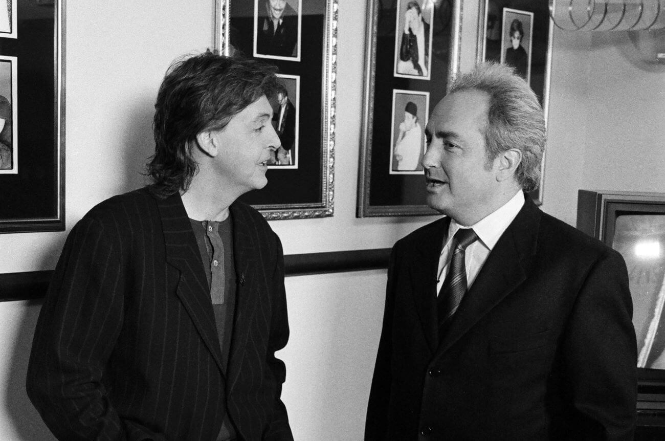 A black and white picture of Paul McCartney speaking with Lorne Michaels in front of a wall of photos.