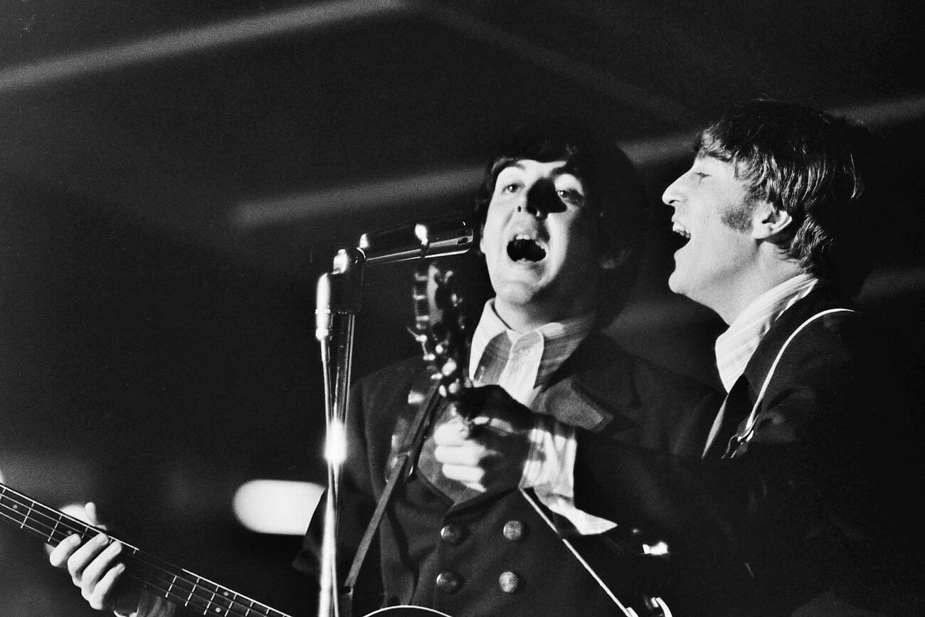 A black and white picture of Paul McCartney and John Lennon playing guitars and singing into the same microphone.