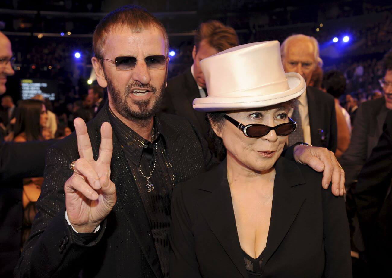 Ringo Starr holds up a peace sign and rests his hand on Yoko Ono's shoulder. She wears sunglasses and a white top hat.