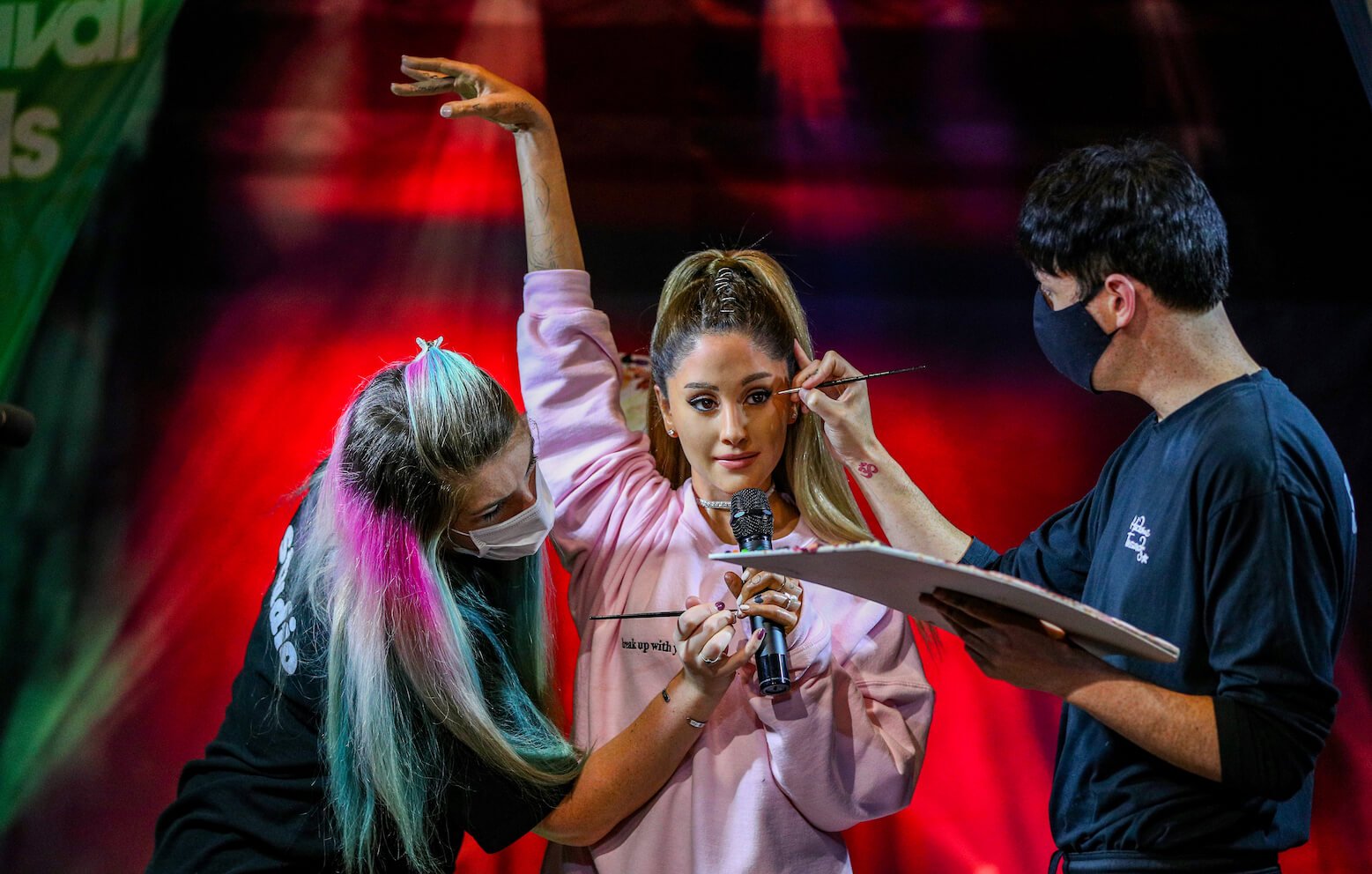 Ariana Grande holding her arm up in a pose while two makeup artists fix her face makeup on stage