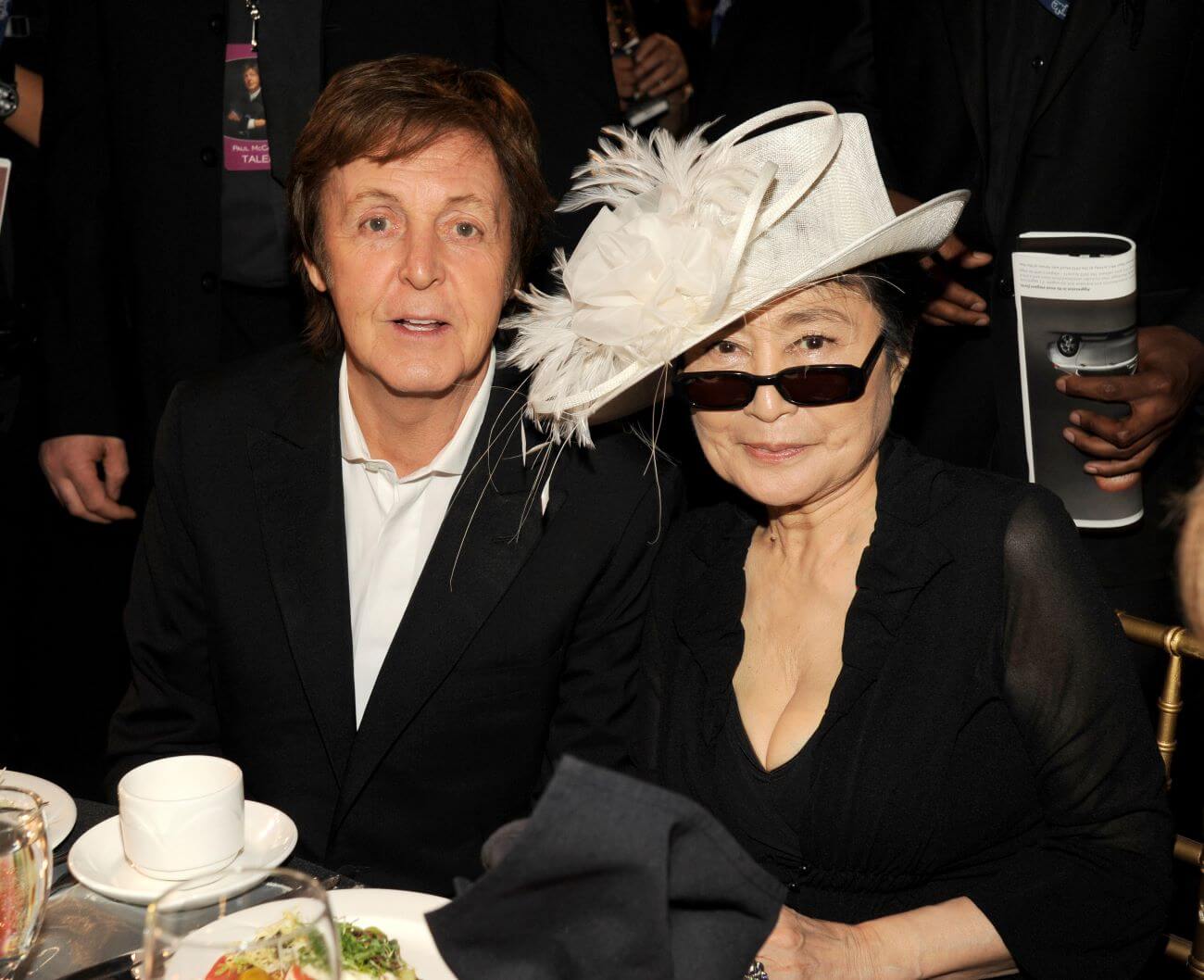 Paul McCartney and Yoko Ono sit at a table together. Ono wears sunglasses and a hat.