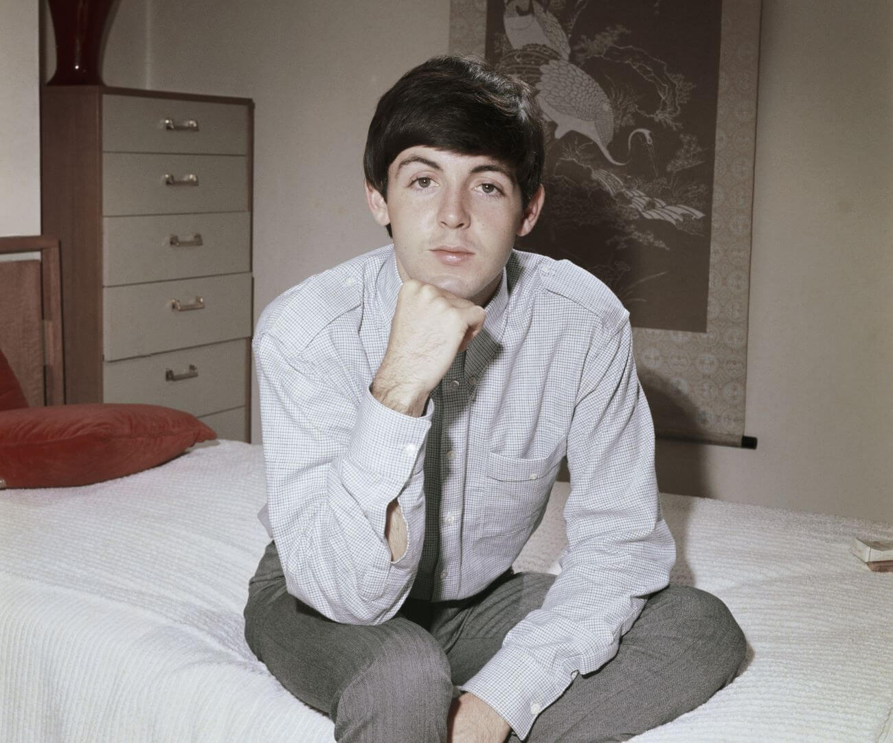 Paul MCCartney sits on the edge of a bed with white sheets. He rests his chin on his fist and his elbow on his thigh.