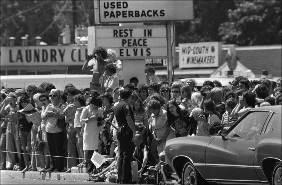 A crowd of people gathers outside a strip mall beneath a sign that says 'Rest in Peace Elvis.'