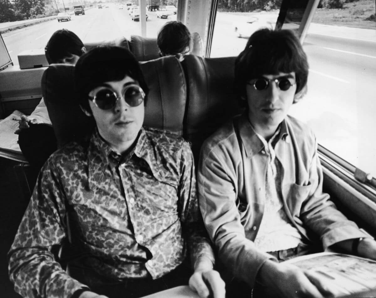 A black and white picture of Paul McCartney and George Harrison wearing sunglasses and sitting on a bus.