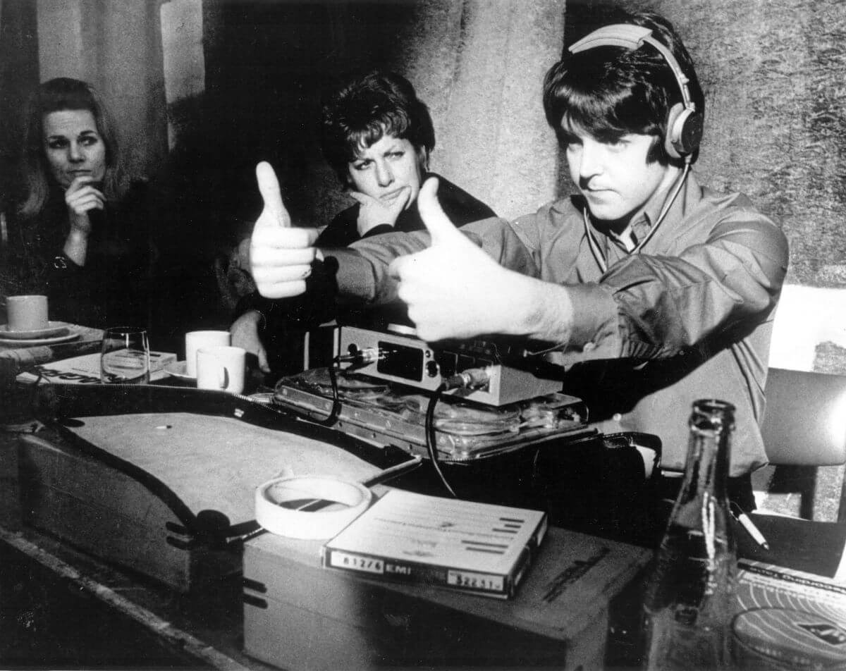 A black and white picture of Paul McCartney wearing headphones and giving two thumbs up while two women look on.