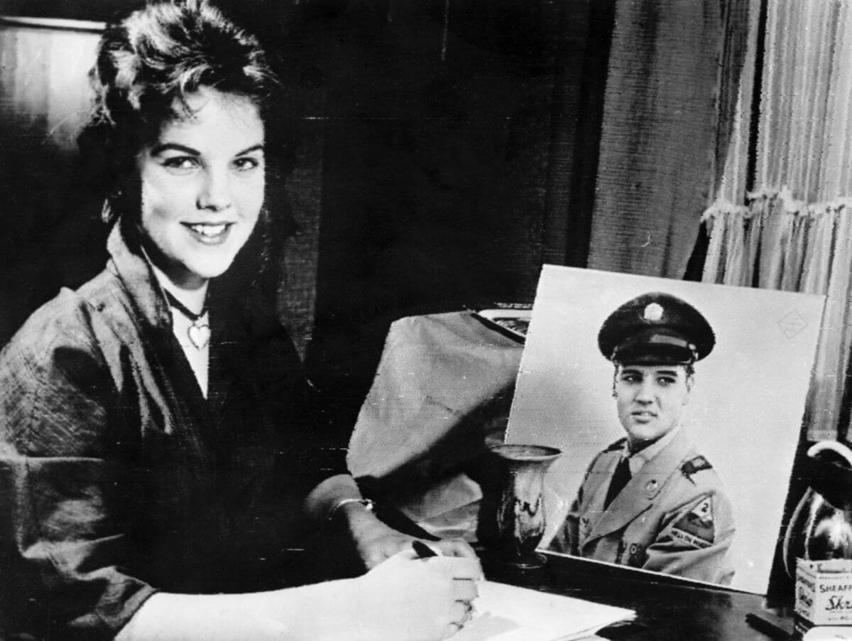 A black and white picture of Priscilla Presley writing a letter while seated before a photo of Elvis in his uniform.