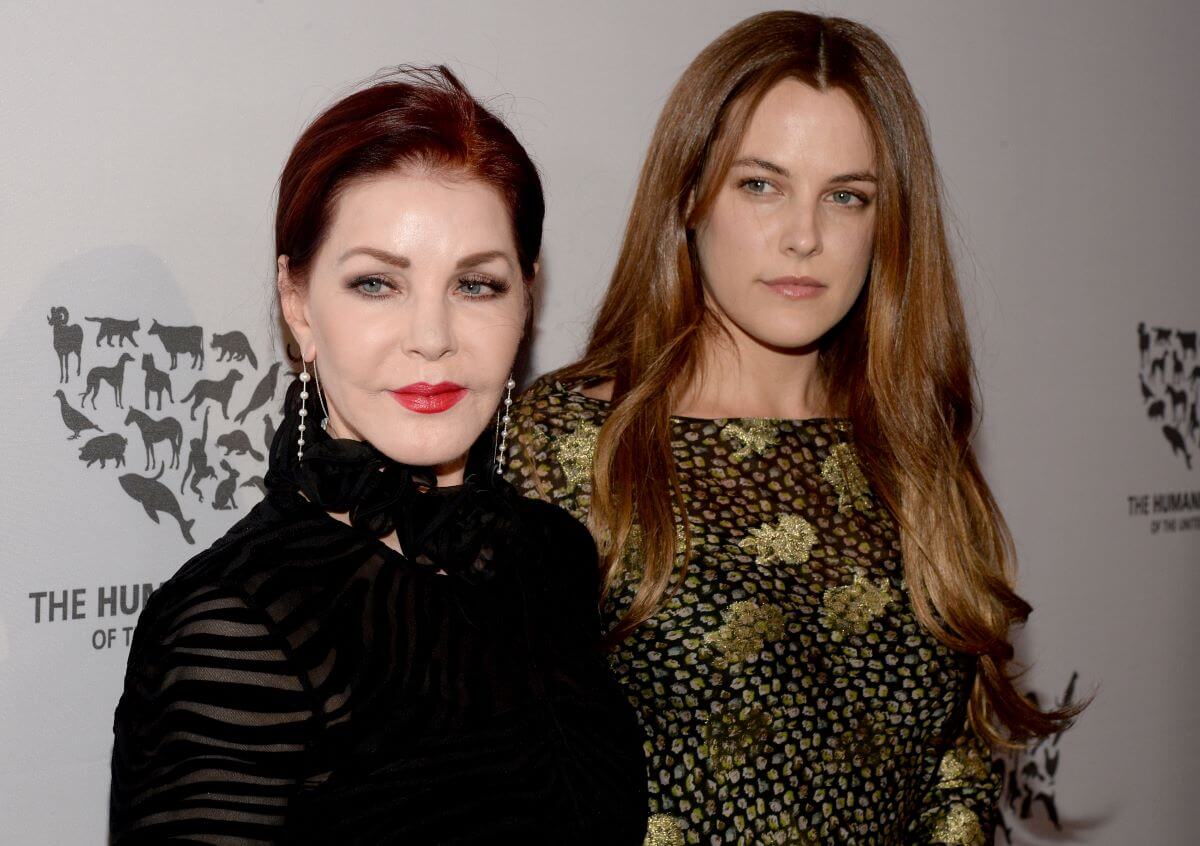 Priscilla Presley wears a black dress and red lipstick. She stands next to Riley Keough, who wears a gold and green dress.
