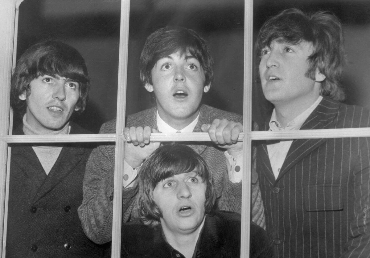 A black and white picture of George Harrison, Paul McCartney, John Lennon, and Ringo Starr looking out a window. Starr crouches below his bandmates and McCartney holds the window frame.