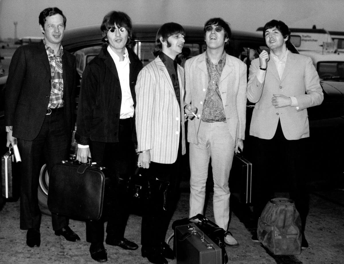 A black and white picture of Brian Epstein, George Harrison, Ringo Starr, John Lennon, and Paul McCartney standing with their bags in front of a car.