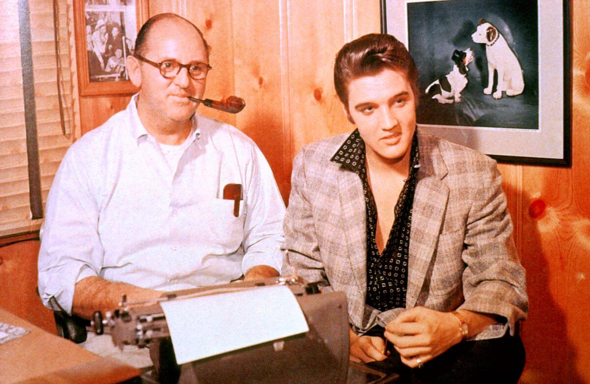 Colonel Tom Parker sits in front of a typewriter and holds a pipe in his mouth. Elvis sits next to him under a painting of two dogs.