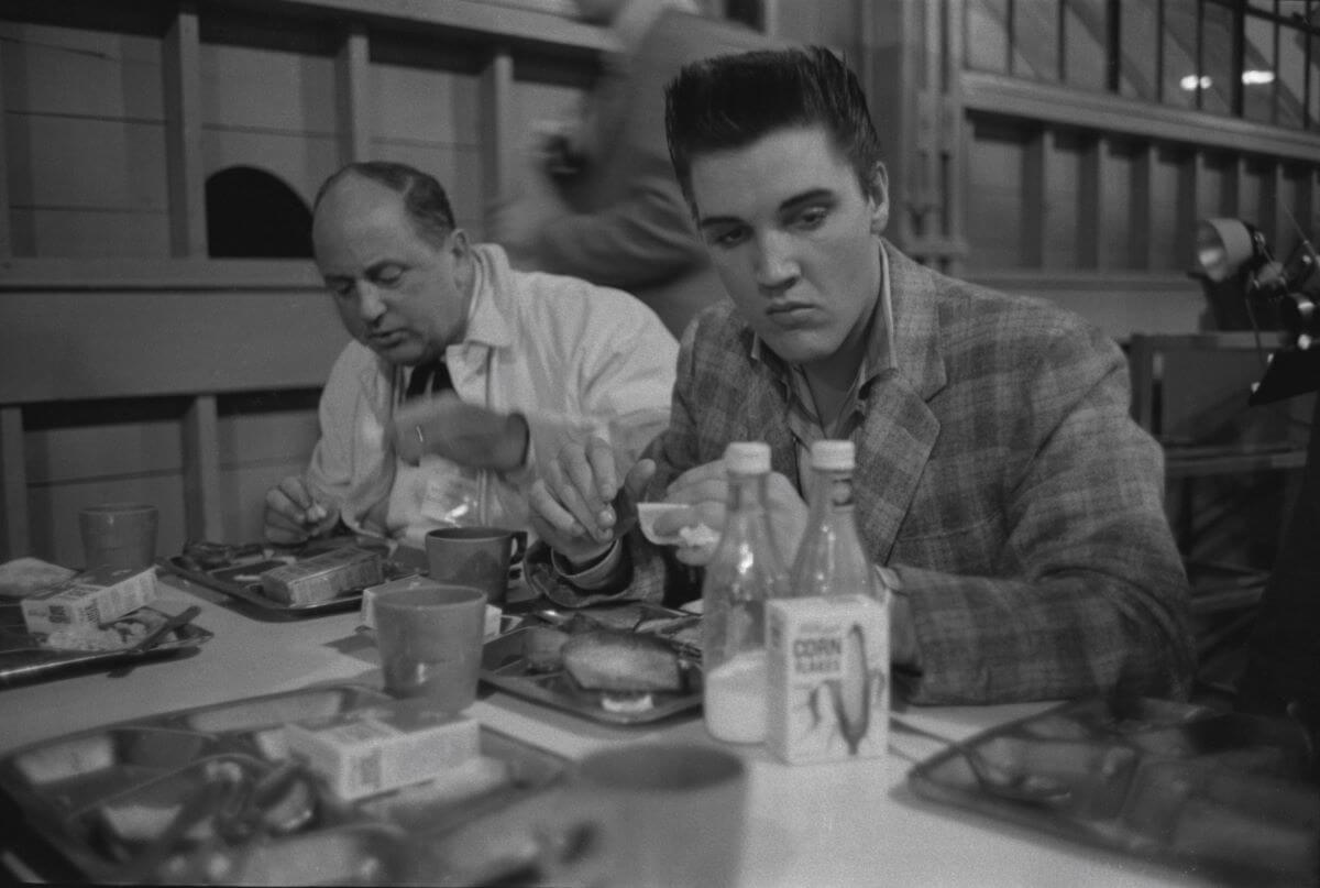 A black and white picture of Elvis Presley eating breakfast next to Colonel Tom Parker.