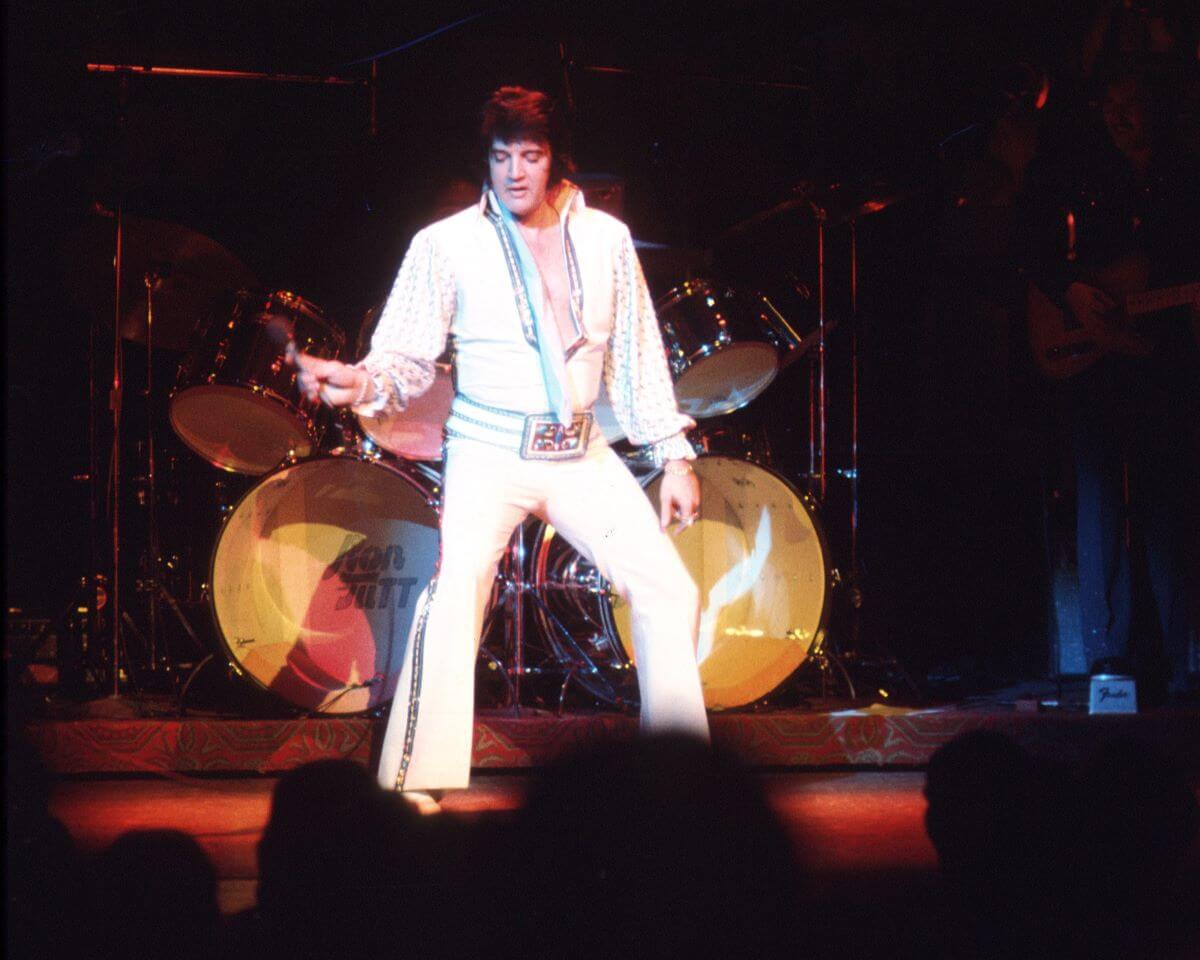 Elvis Presley stands onstage in a white jumpsuit. 