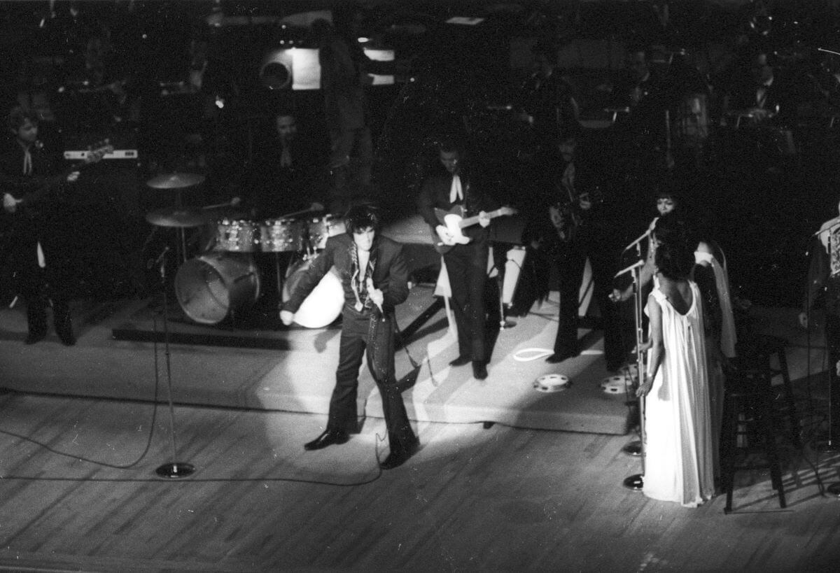 A black and white picture of Elvis Presley singing in a spotlight onstage. His backup singers, the Sweet Inspirations, stand to the side of the stage.