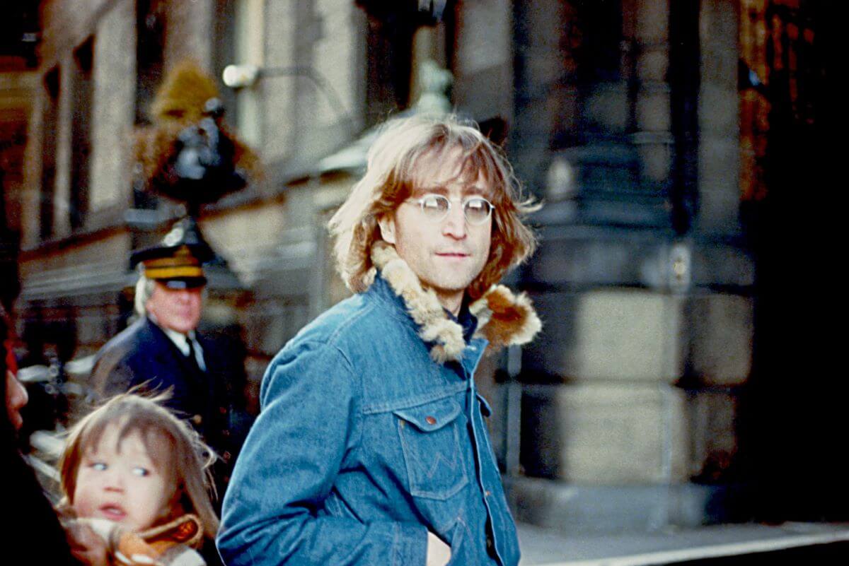 John Lennon wears a denim coat with a fur collar and walks outside.
