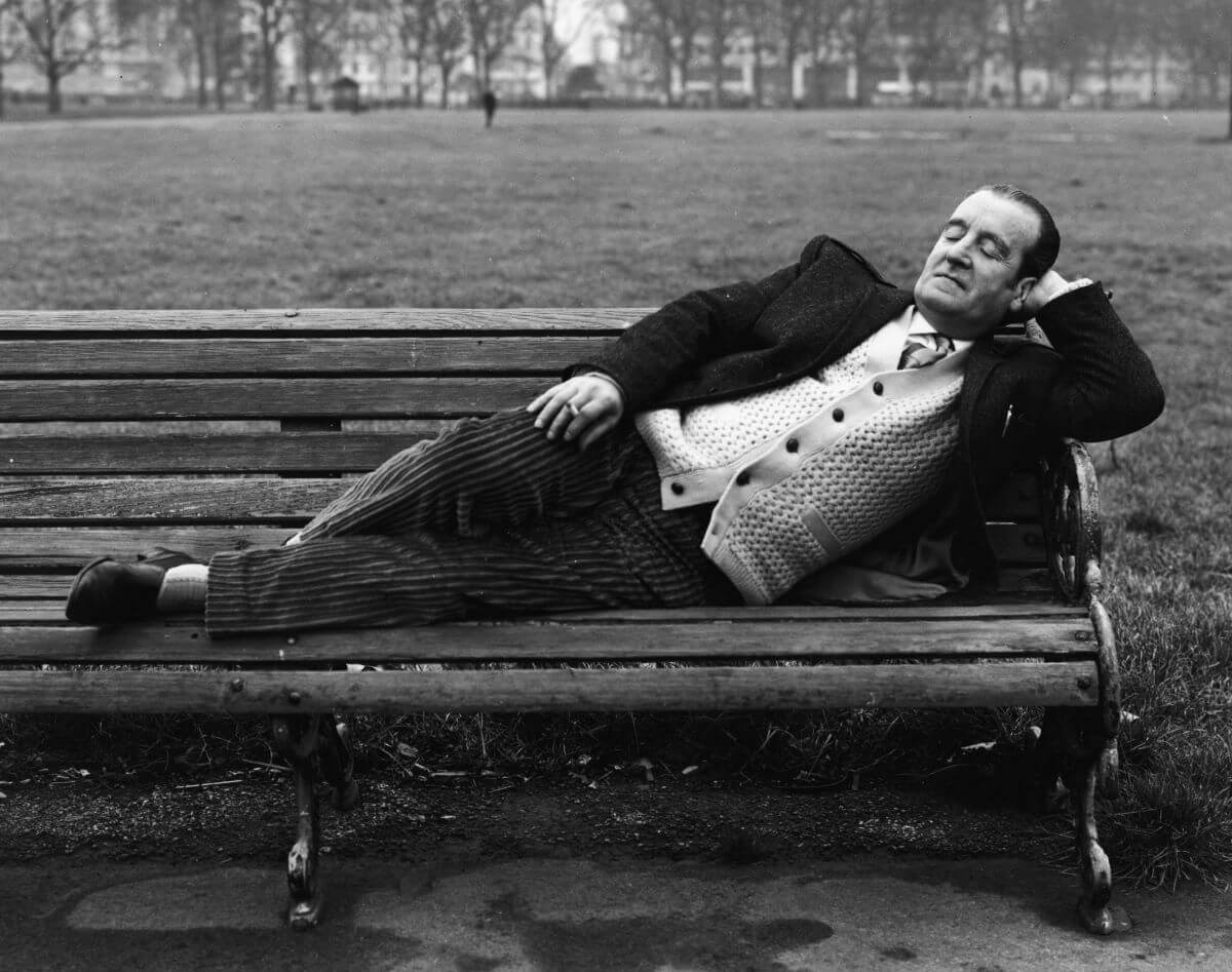 A black and white picture of Alfred Lennon laying on a bench and holding a cigarette in one hand.