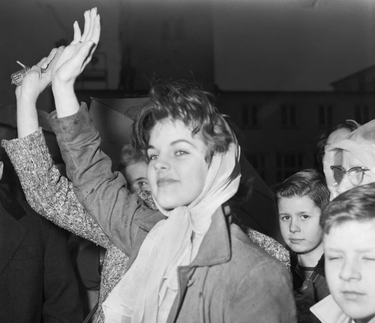 A black and white picture of Priscilla Presley lifting her arm in a wave. She wears a scarf around her hair and stands in a crowd.