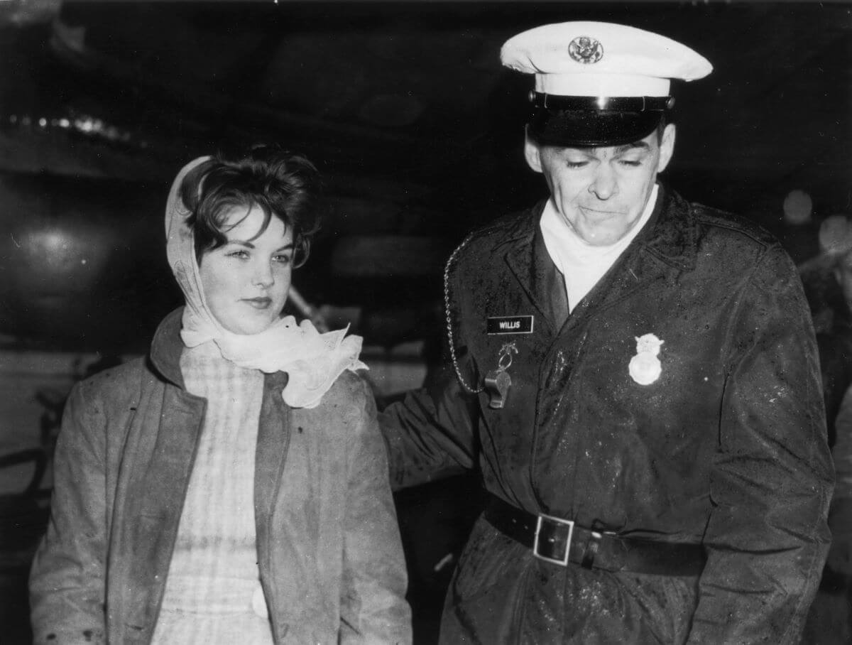 A black and white picture of Priscilla Presley walking with a police officer. She wears a jacket and a scarf in her hair.