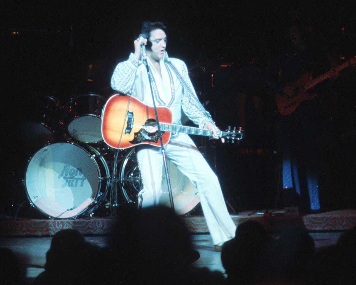 Elvis Presley wears a white jumpsuit and holds a microphone and acoustic guitar.