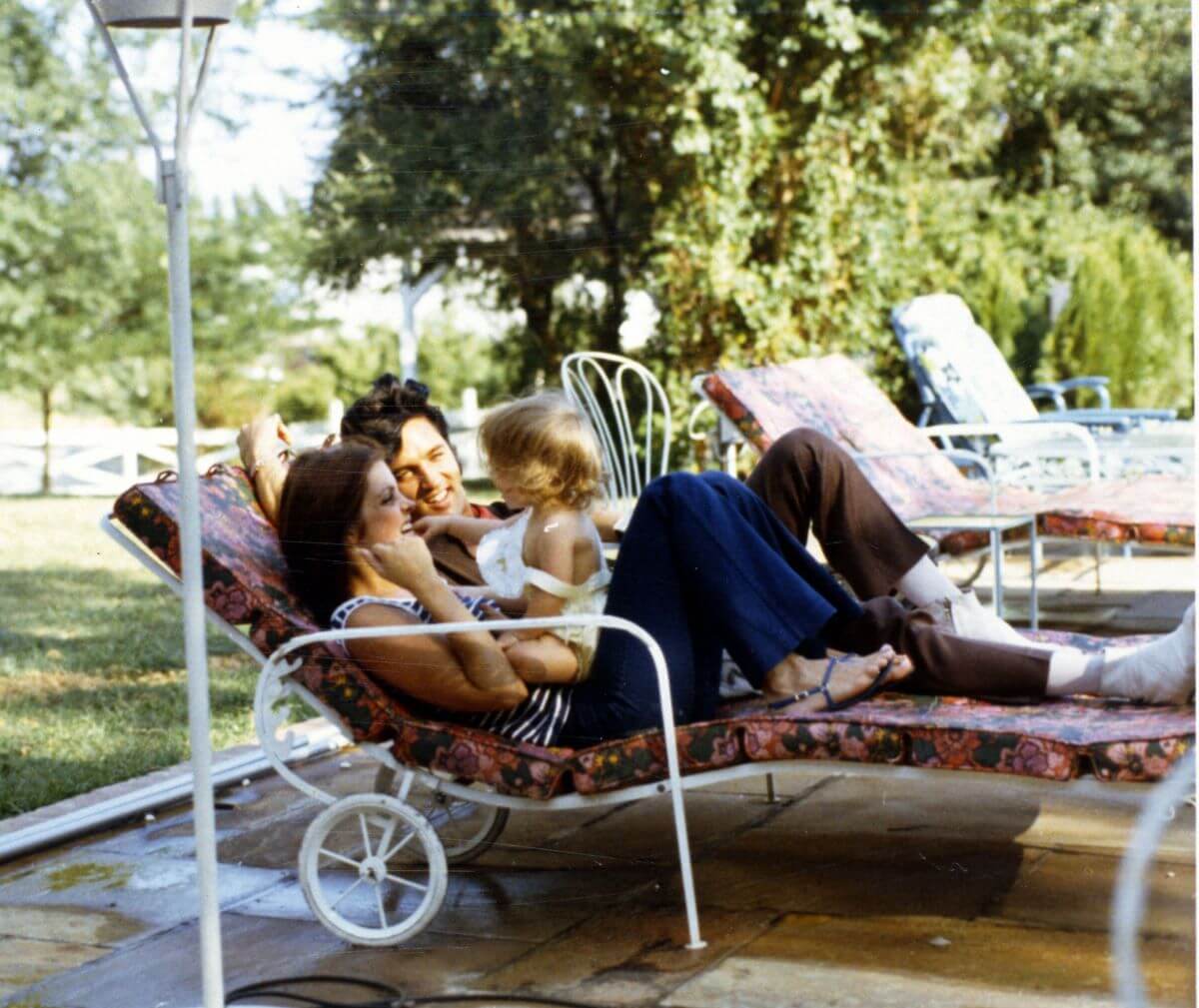 Priscilla and Elvis Presley lay in lounge chairs while Lisa Marie Presley sits on Priscilla's stomach.