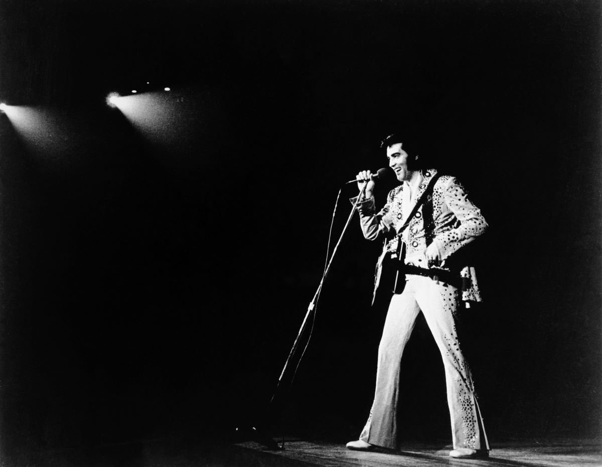 Elvis Presley stands on a stage and holds a microphone. He has a guitar strapped to his body.