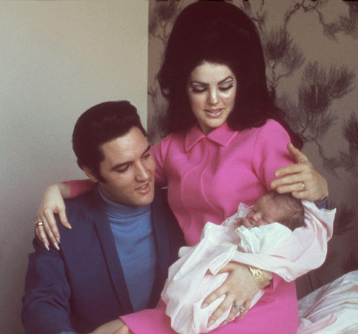 Priscilla Presley sits on Elvis' lap and holds an infant Lisa Marie Presley. Elvis' arm is around her.