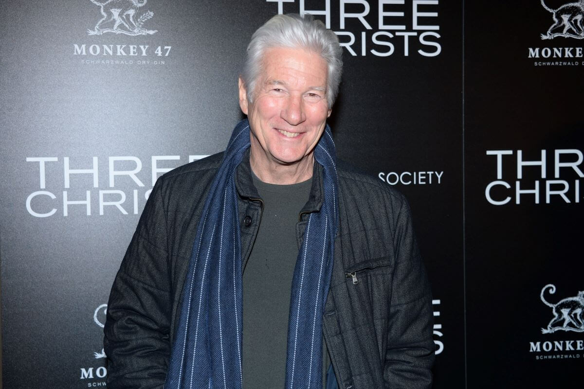 Richard Gere wears a jacket and blue scarf while standing against a black background.