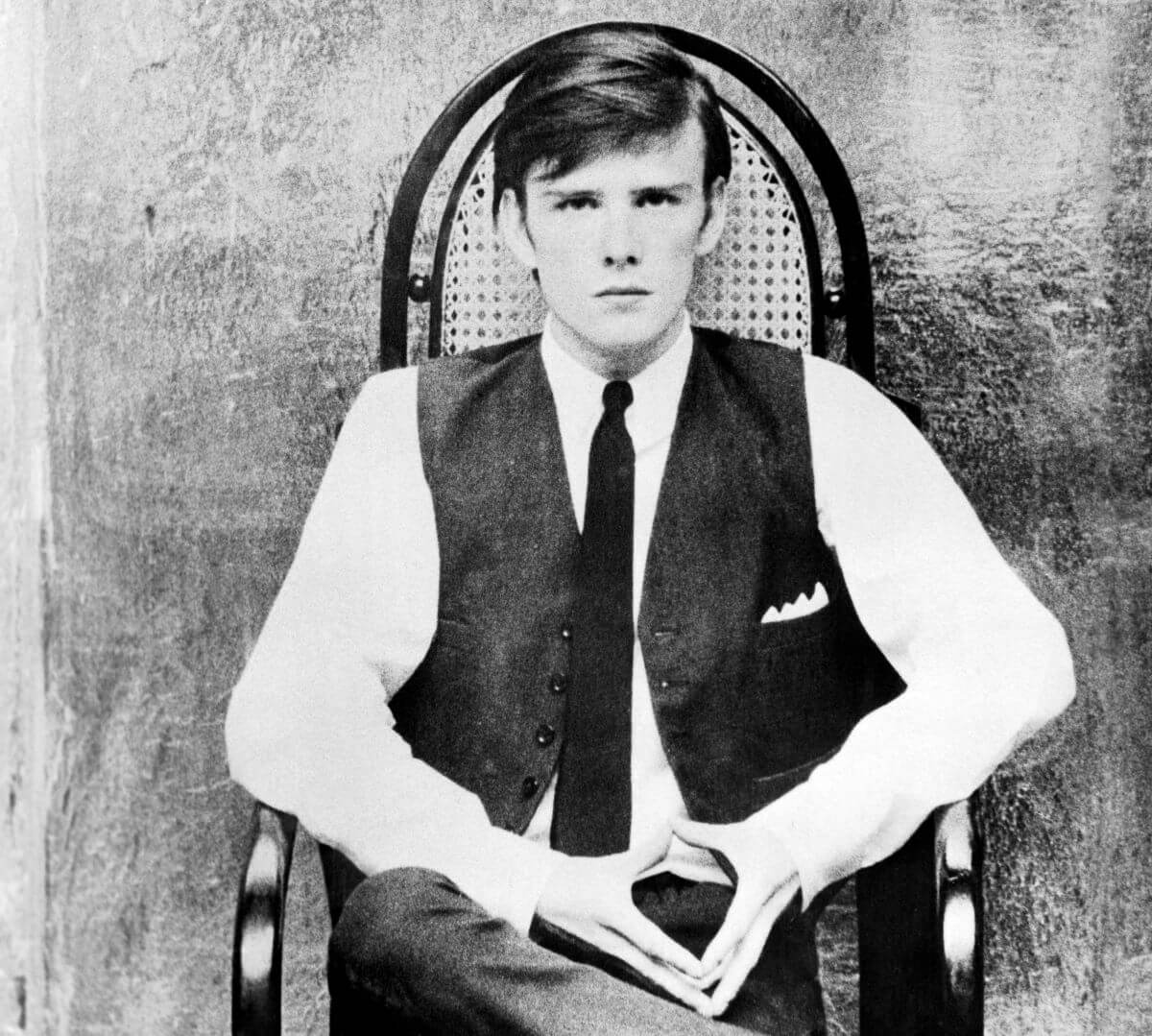 A black and white picture of Stuart Sutcliffe wearing a tie and sitting in a chair.