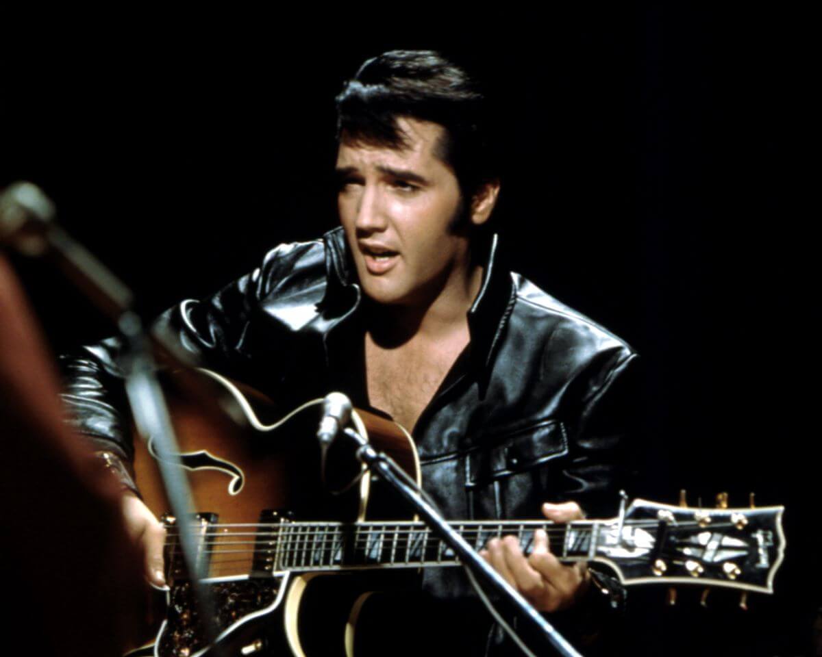 Elvis Presley wears a leather jacket and sits with a guitar.