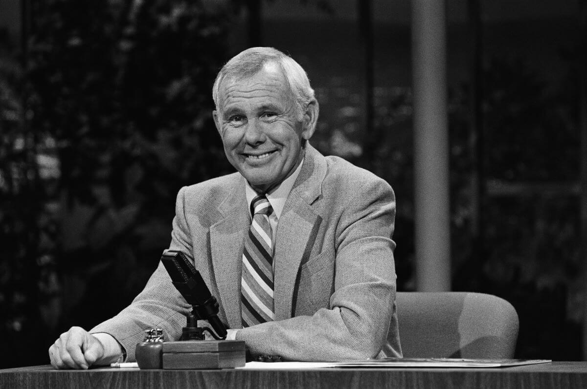 A black and white picture of Johnny Carson sitting at a desk with a microphone in front of him.